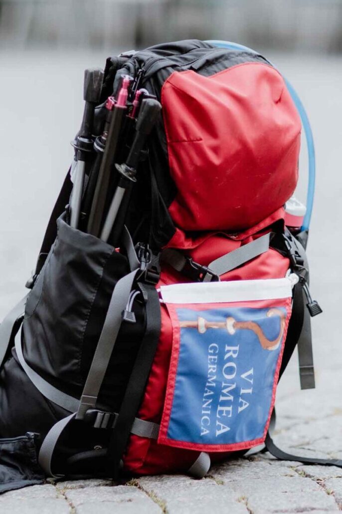 Ein Rucksack mit einer Fahne auf der das logo der Via Romea zu sehen ist, steht auf einem geplastertem Platz.